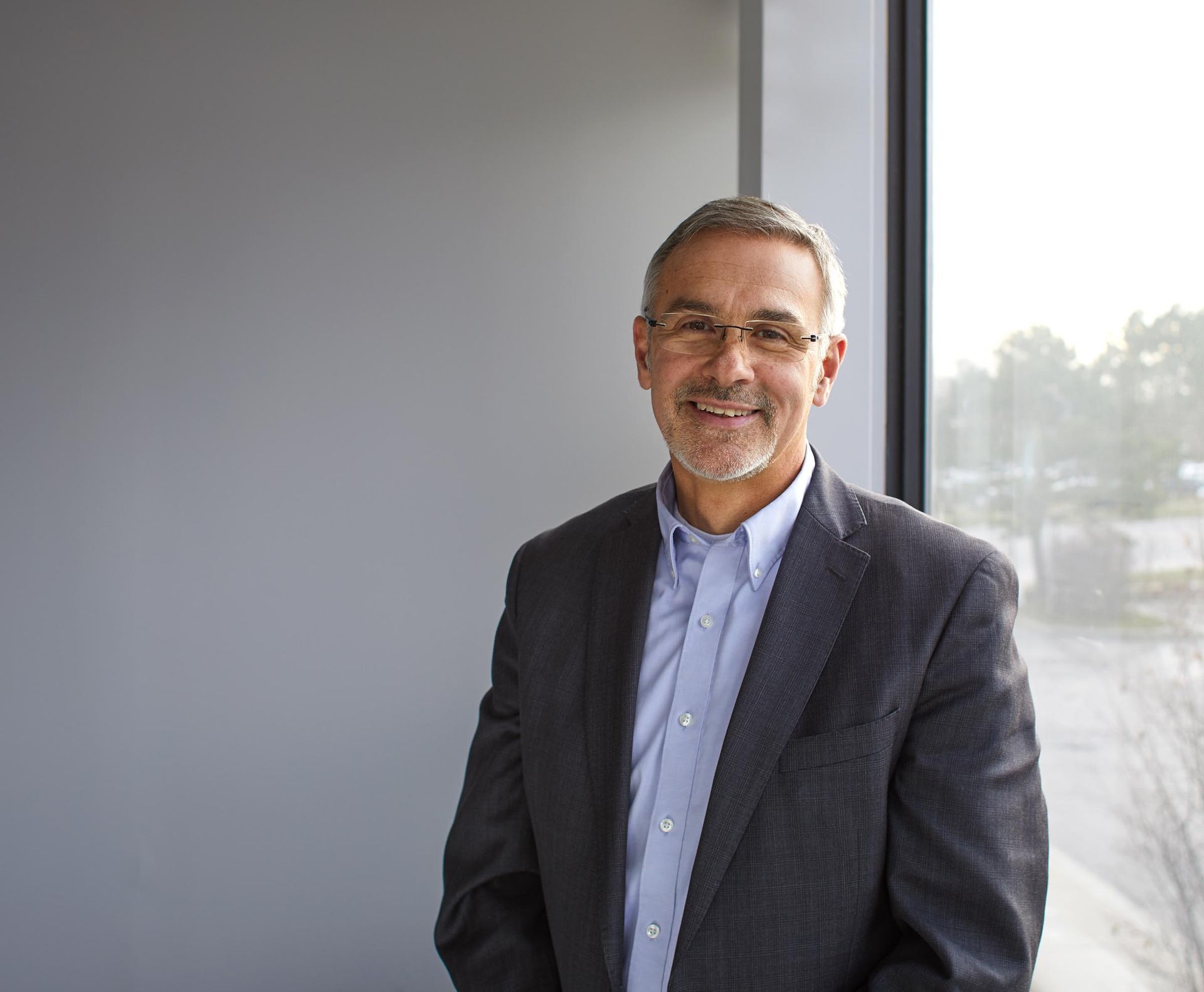 A man in a suit and glasses is standing in front of a window.