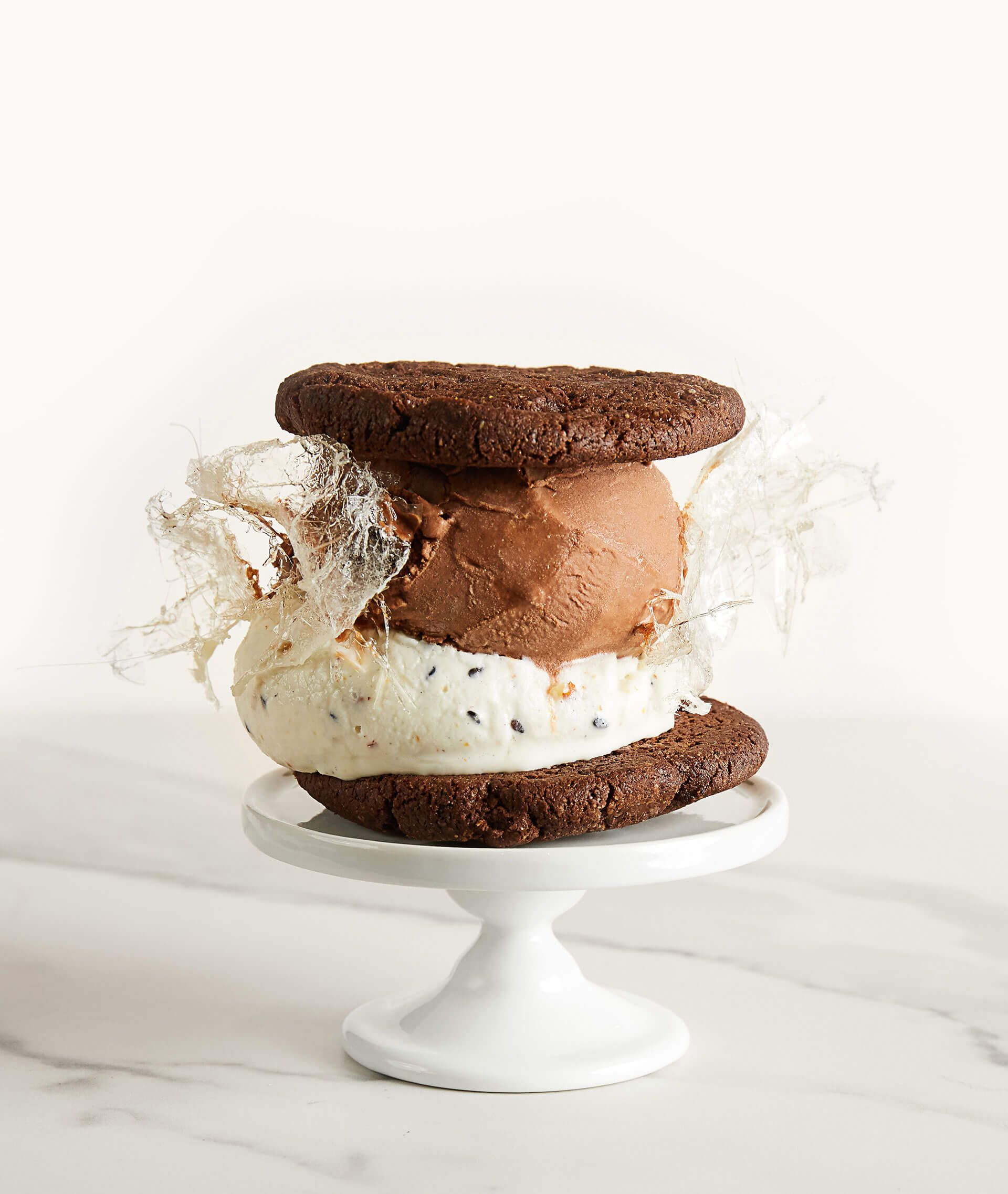 A chocolate cookie ice cream sandwich is sitting on top of a white cake stand.