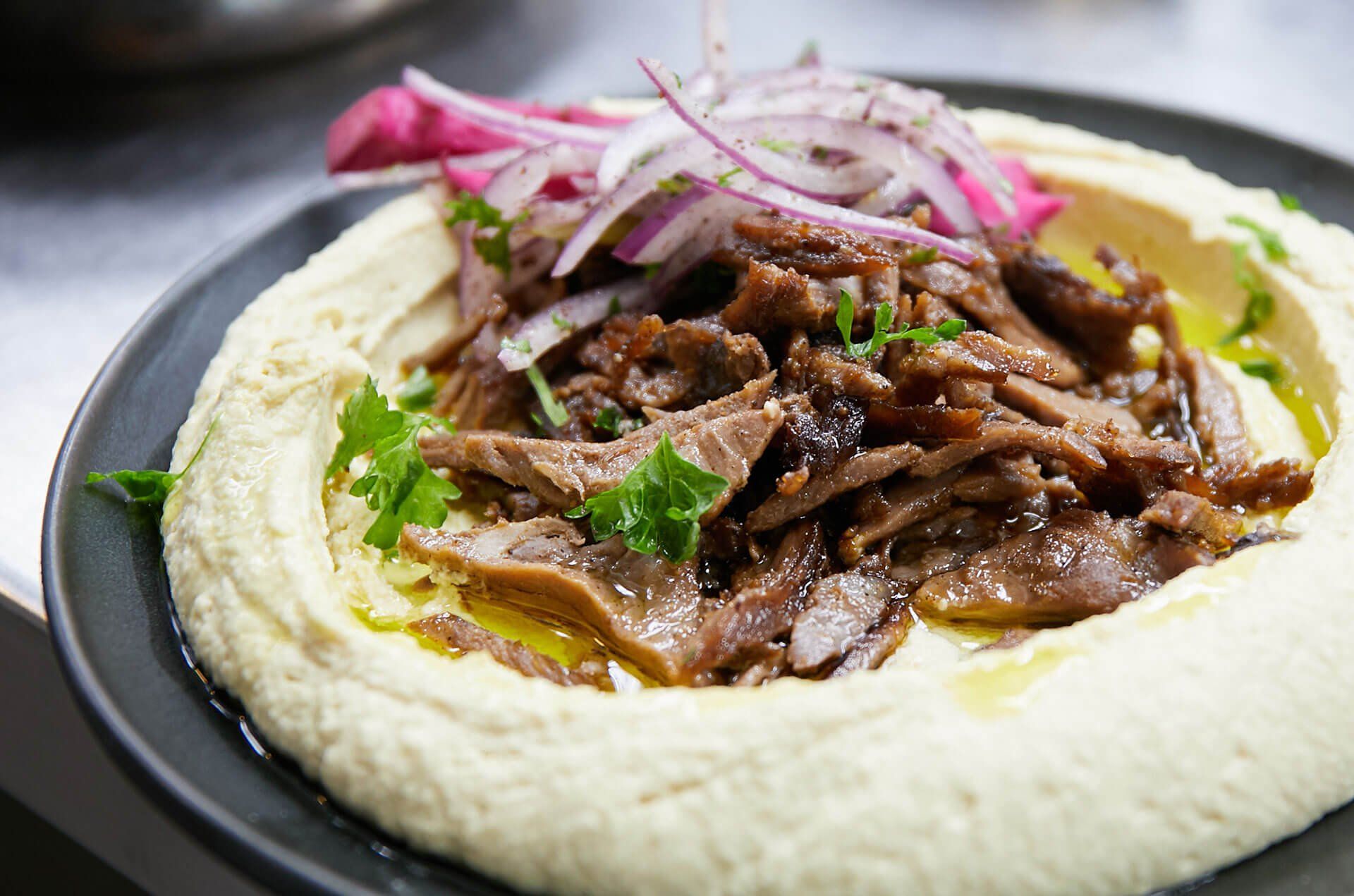 A close up of a plate of hummus with meat and onions on a table.
