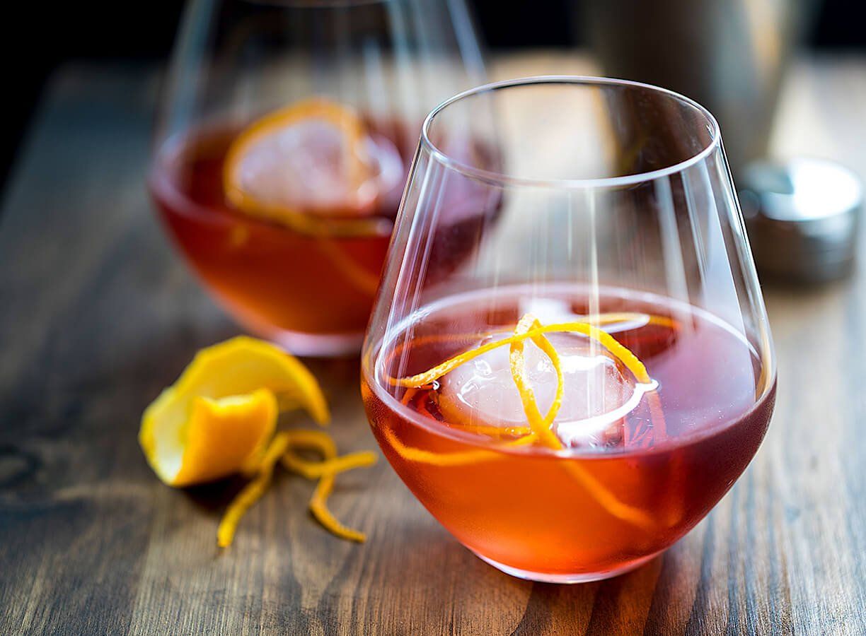 Two glasses of alcohol with ice and orange peel on a wooden table.