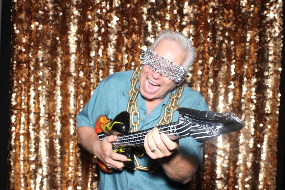 A man is holding an inflatable guitar in front of a gold sequined backdrop.