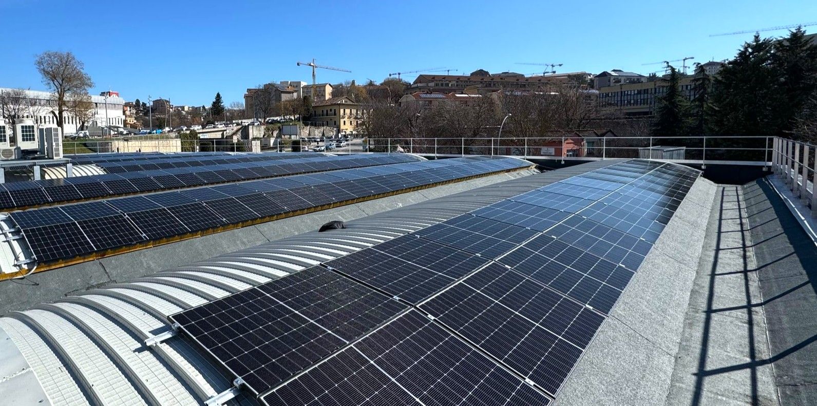 Pannelli solari sul tetto di un edificio con un cielo azzurro sullo sfondo. Altri edifici e alberi sono visibili in lontananza.