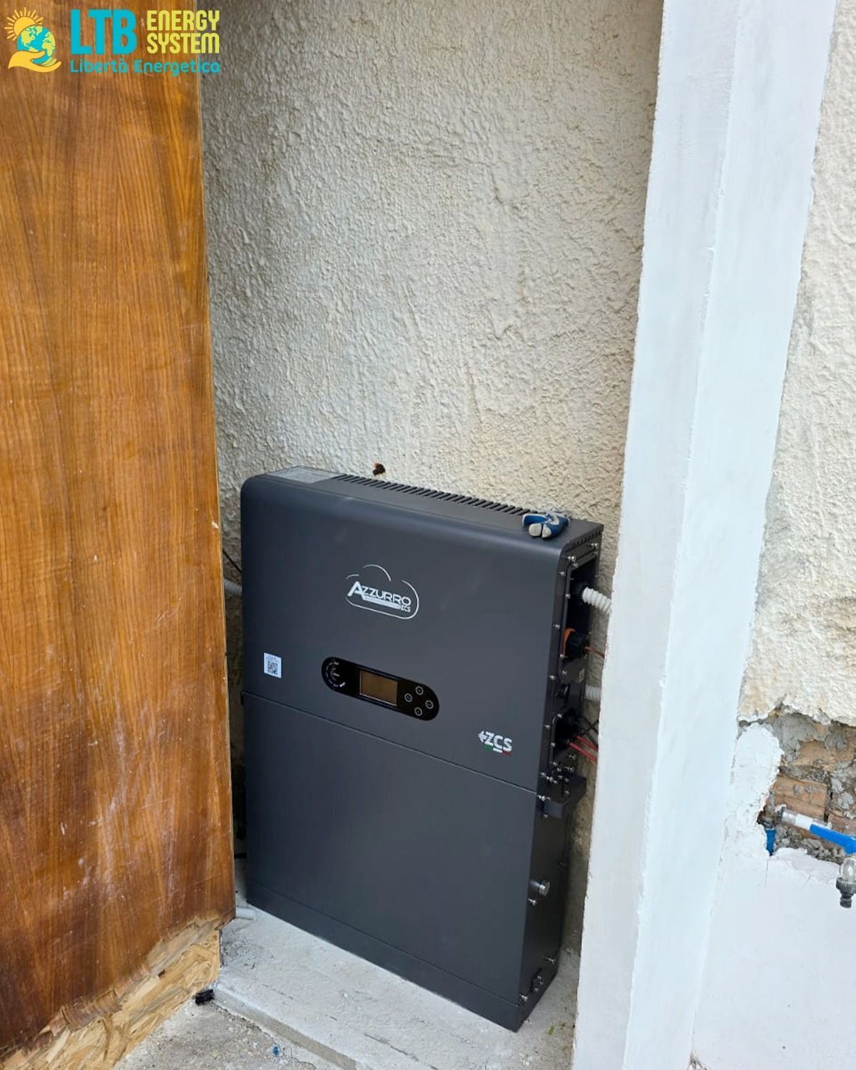 Un inverter solare grigio scuro installato all'esterno, tra una porta di legno e una parete in stucco. L'inverter è dotato di un display e di diversi punti di connessione.