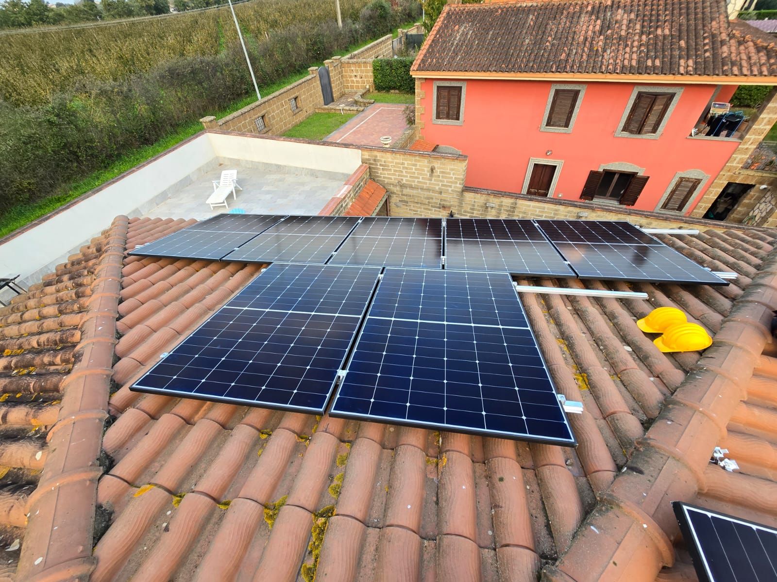 Pannelli solari installati sul tetto di tegole di terracotta di un edificio con facciata rossa; sul tetto è presente un casco giallo.