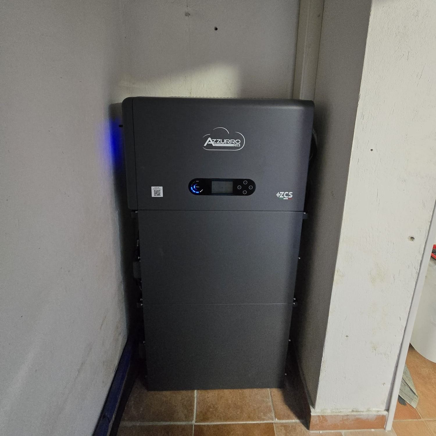 Un'unità di batteria solare grigia installata in un angolo di una stanza con pareti bianche e pavimento piastrellato. La batteria ha una luce blu sul lato sinistro.