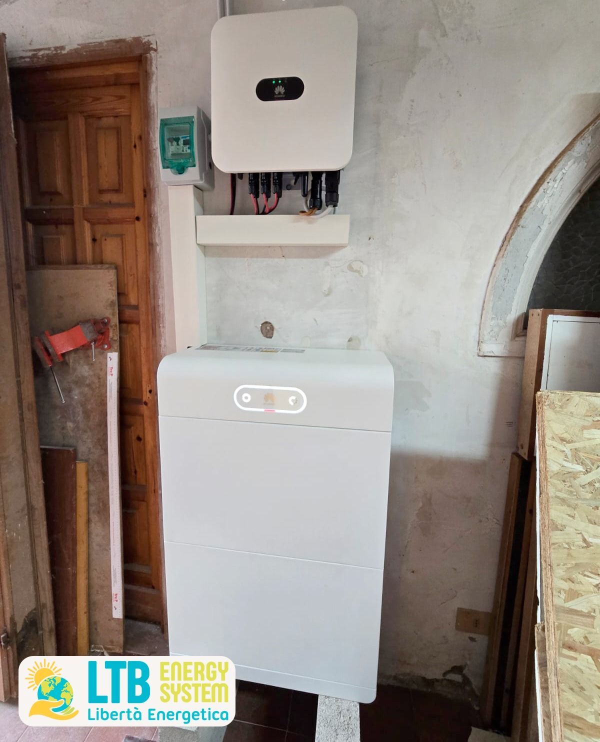 Impianto di pannelli solari installato all'interno, con scatola inverter bianca e batteria contro una parete in legno.