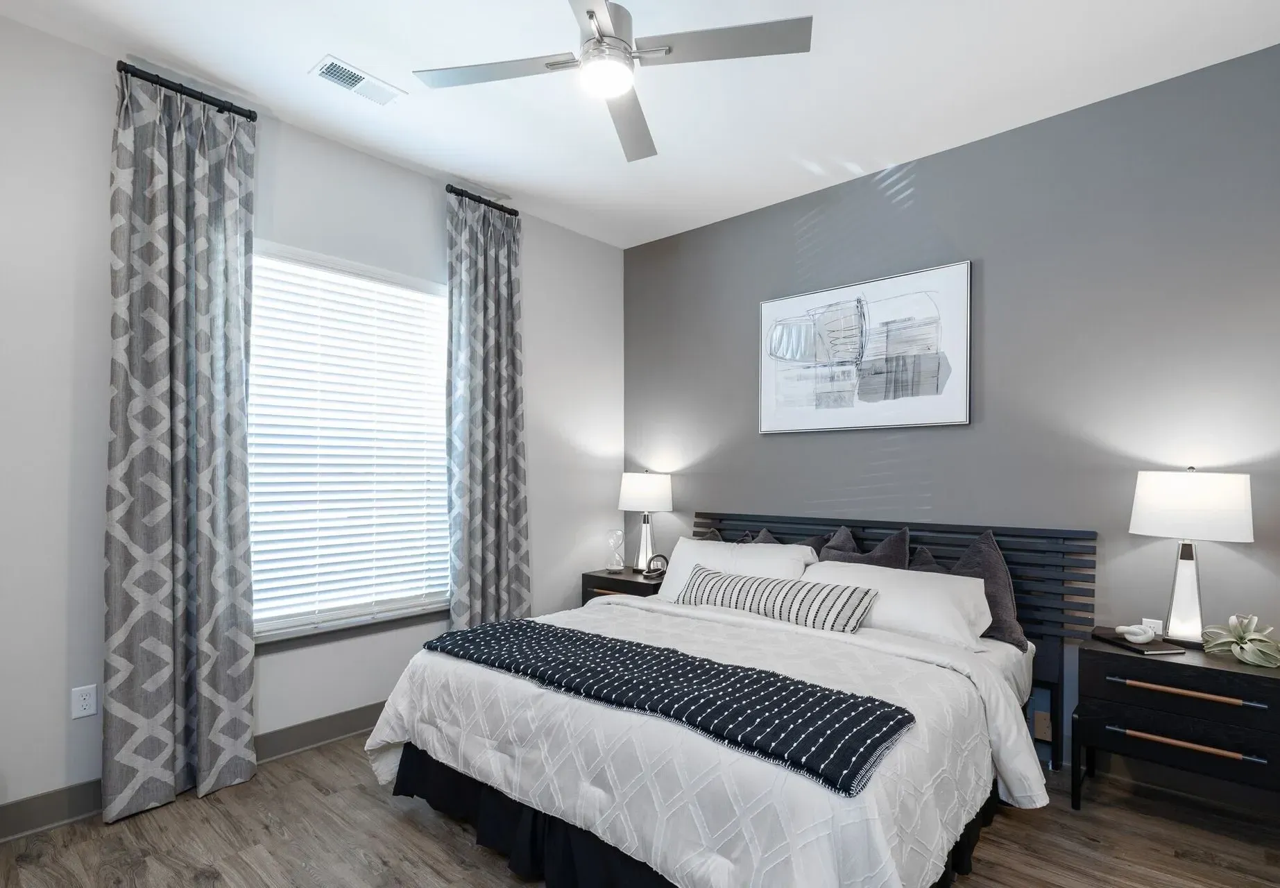 Modern apartment bedroom with a king bed, gray accent wall, two nightstands, and a ceiling fan.