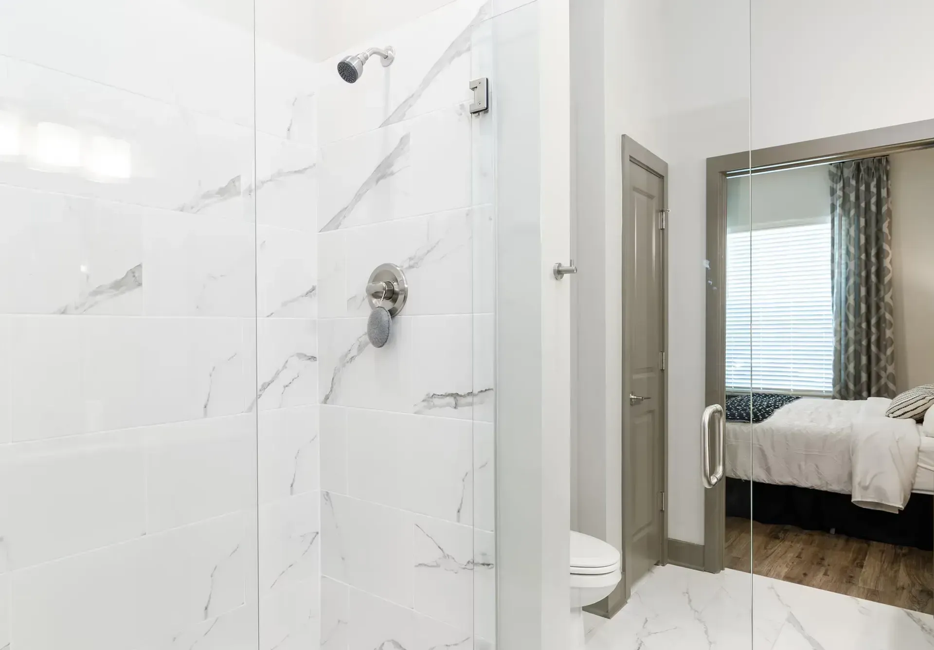 White bathroom with marble-look tiles, glass shower enclosure, and a doorway to a bedroom.