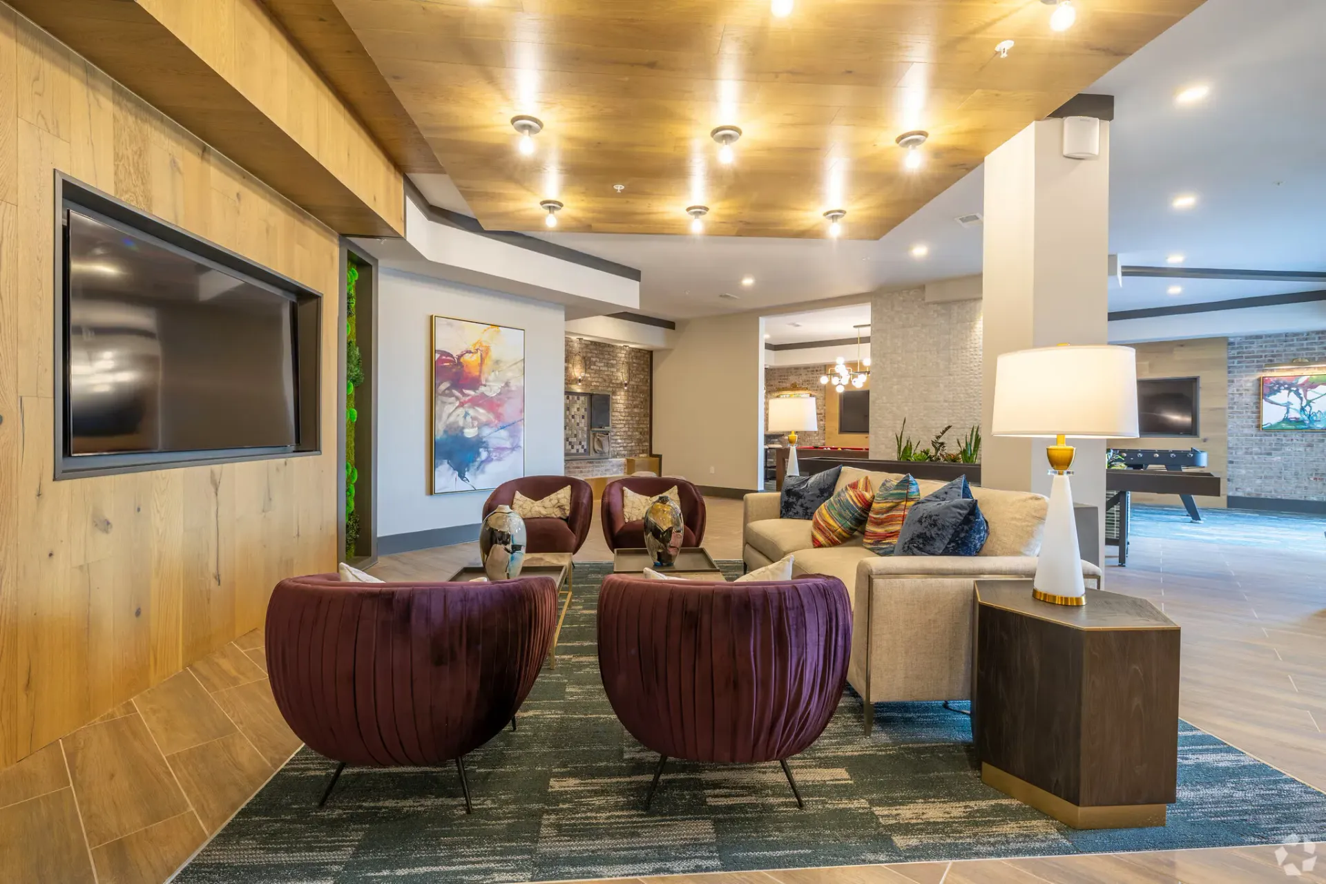 Indoor community lounge with burgundy accent chairs, a beige sofa, and a wall-mounted TV.