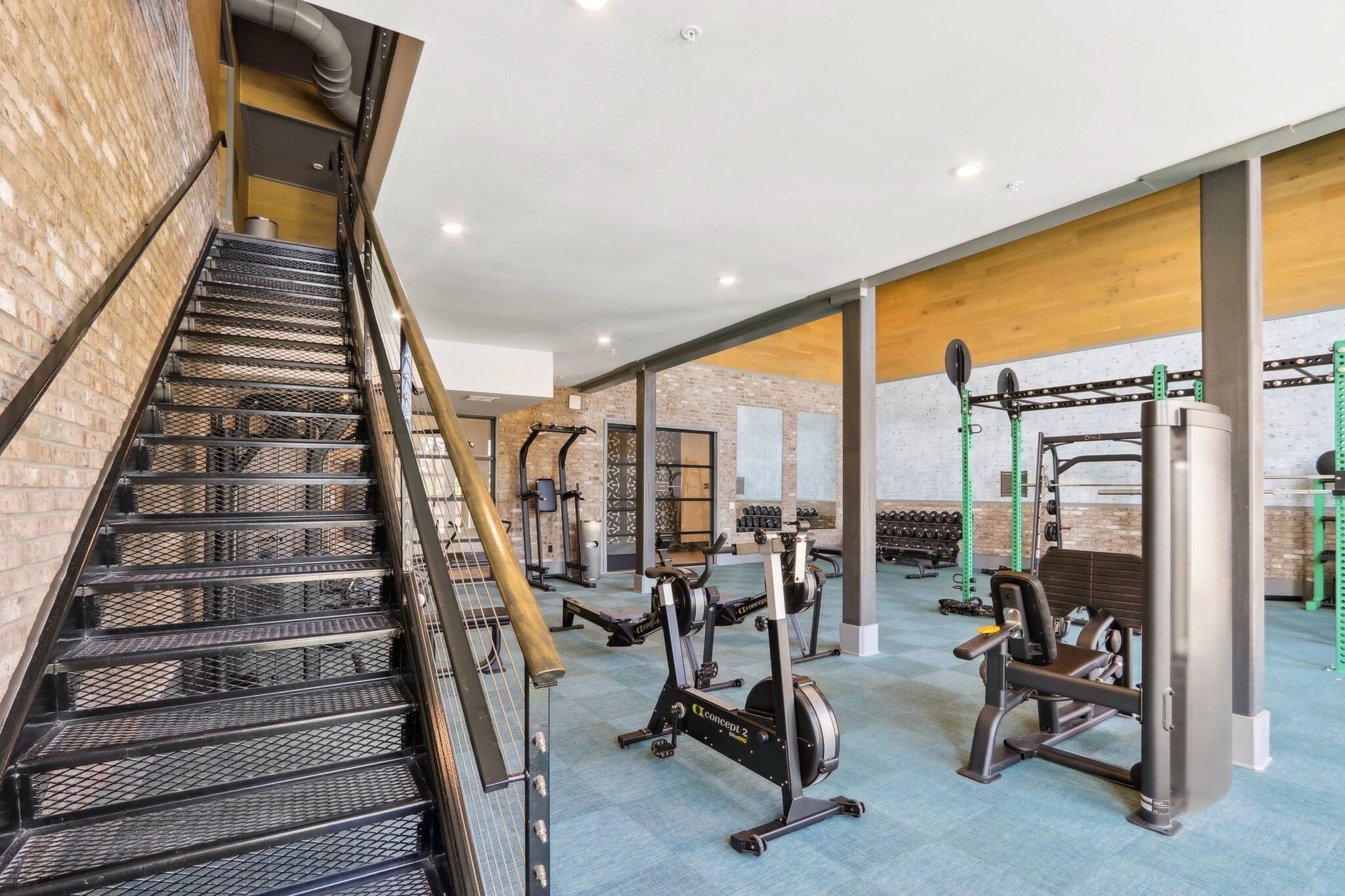 Indoor community gym with cardio machines, free weights, and a metal staircase along brick walls.