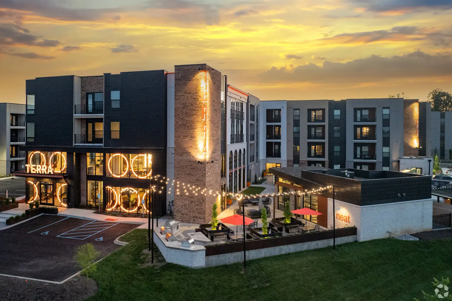 Aerial view of a modern apartment complex at sunset, with courtyard seating, string lights, and a neon Terra sign.