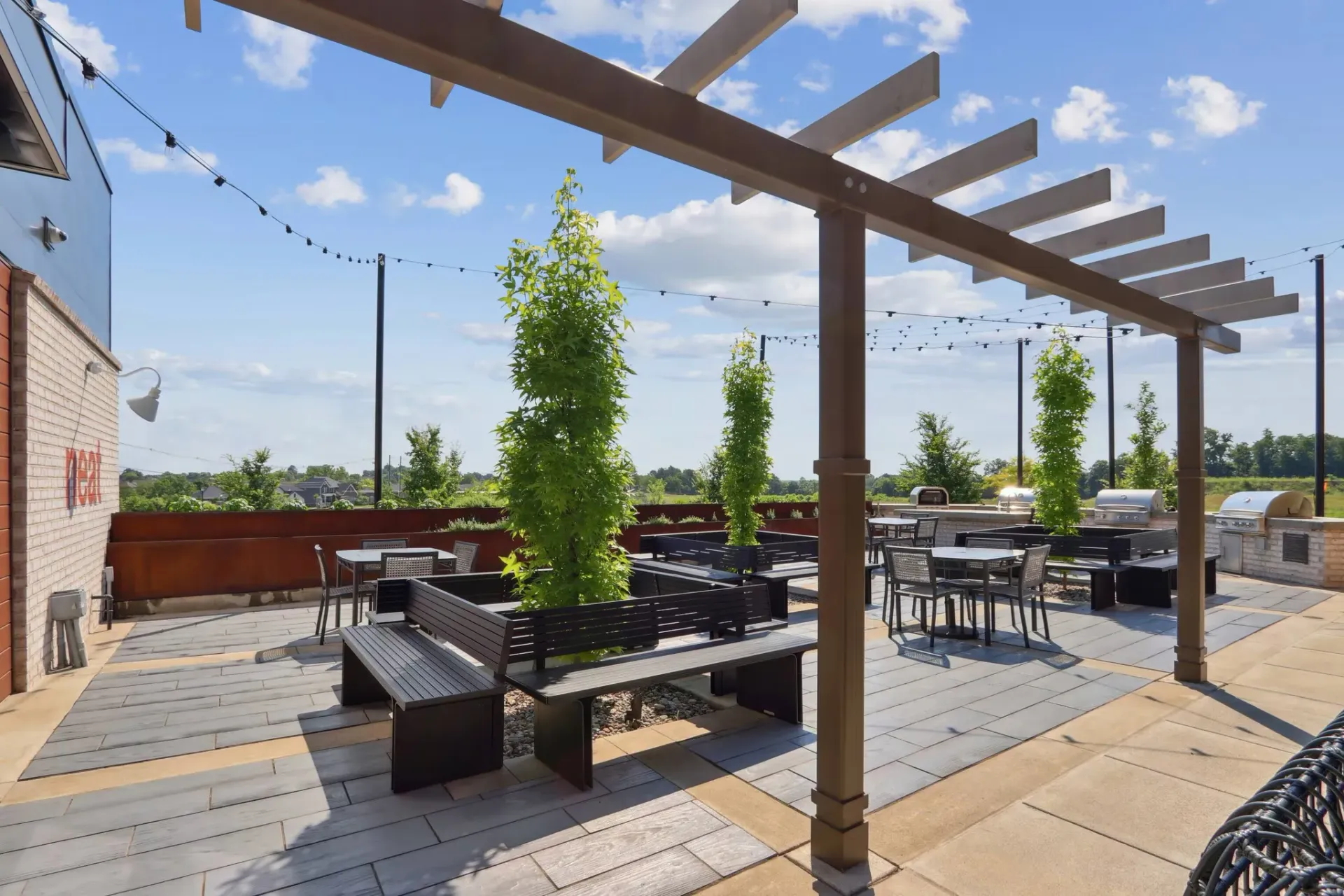 Rooftop community patio under a wooden pergola with string lights, benches, tables, and greenery.