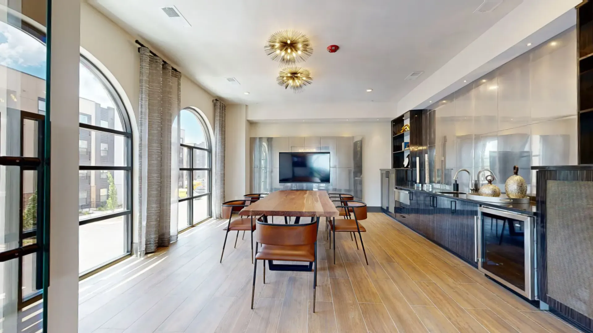 Modern resident lounge with a long wooden table, brown chairs, arched windows, and a sleek kitchenette.