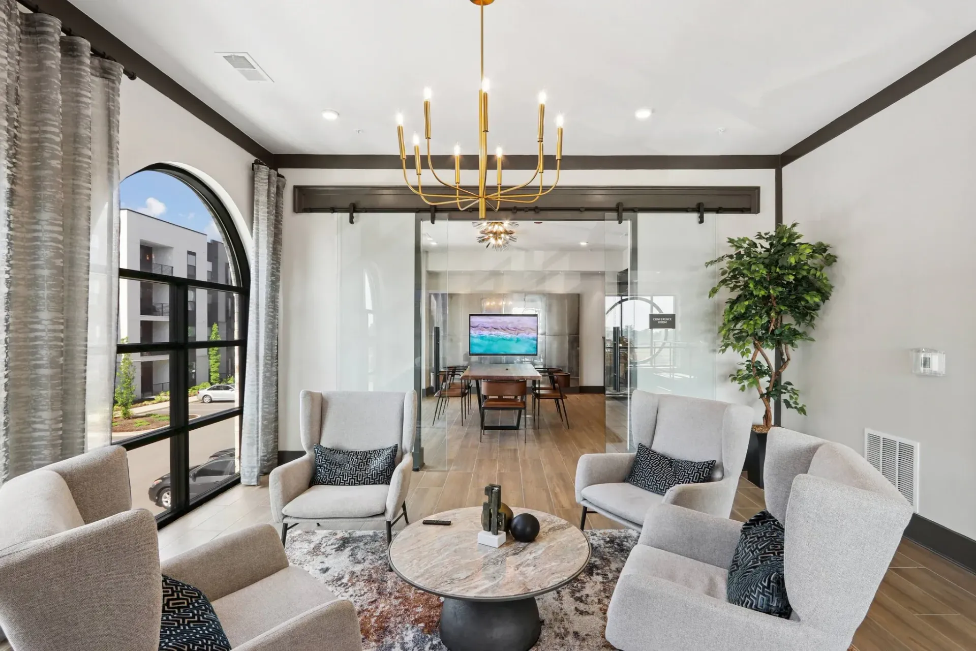 Modern apartment community lounge with arched window, glass doors, chandelier, and gray seating around a marble coffee table.