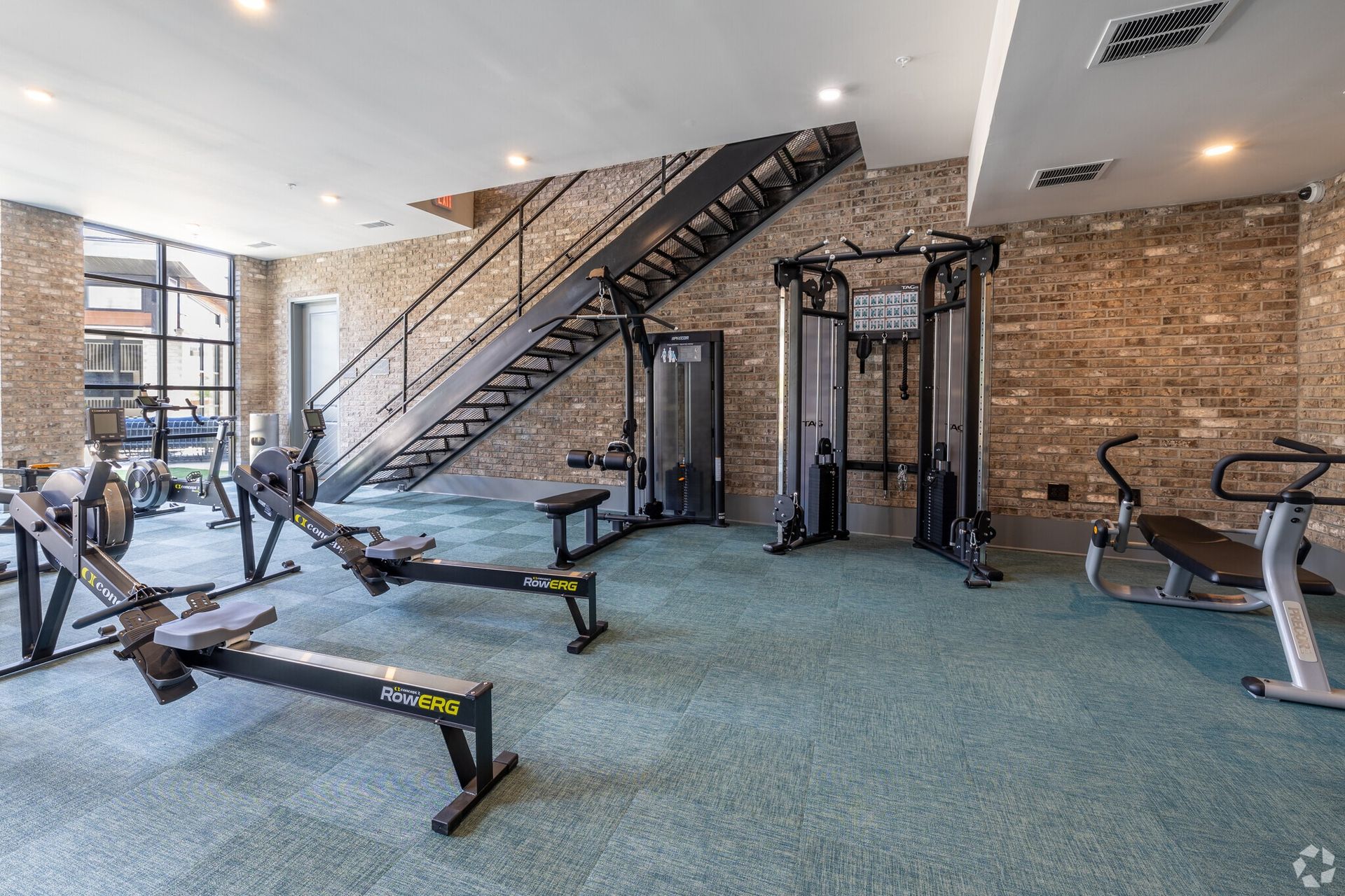 Indoor apartment gym with cardio machines, strength machines, and a metal staircase along brick walls.