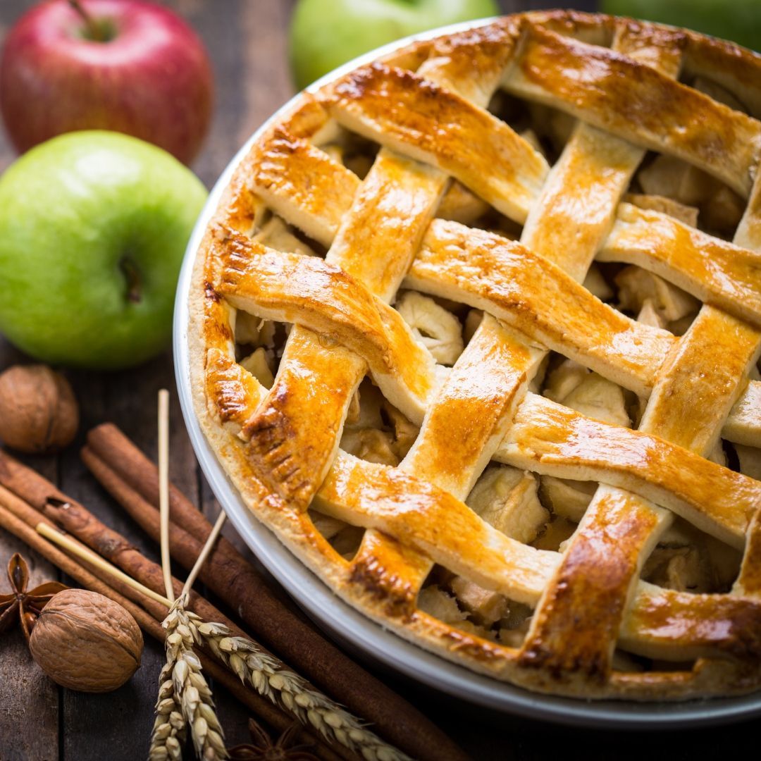 A pie with a lattice crust is on a table with apples and cinnamon sticks