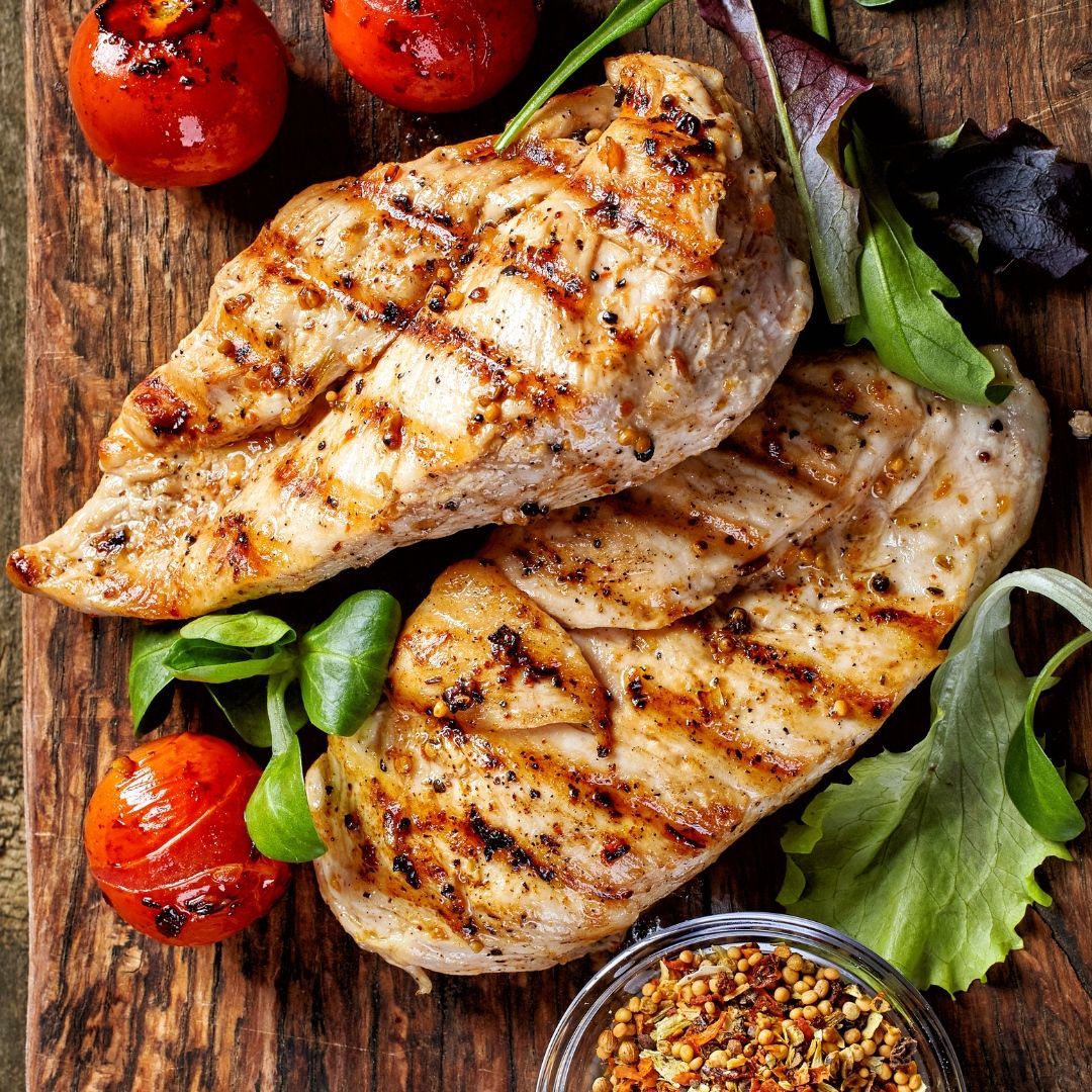 Two grilled chicken breasts on a wooden cutting board with tomatoes and lettuce.