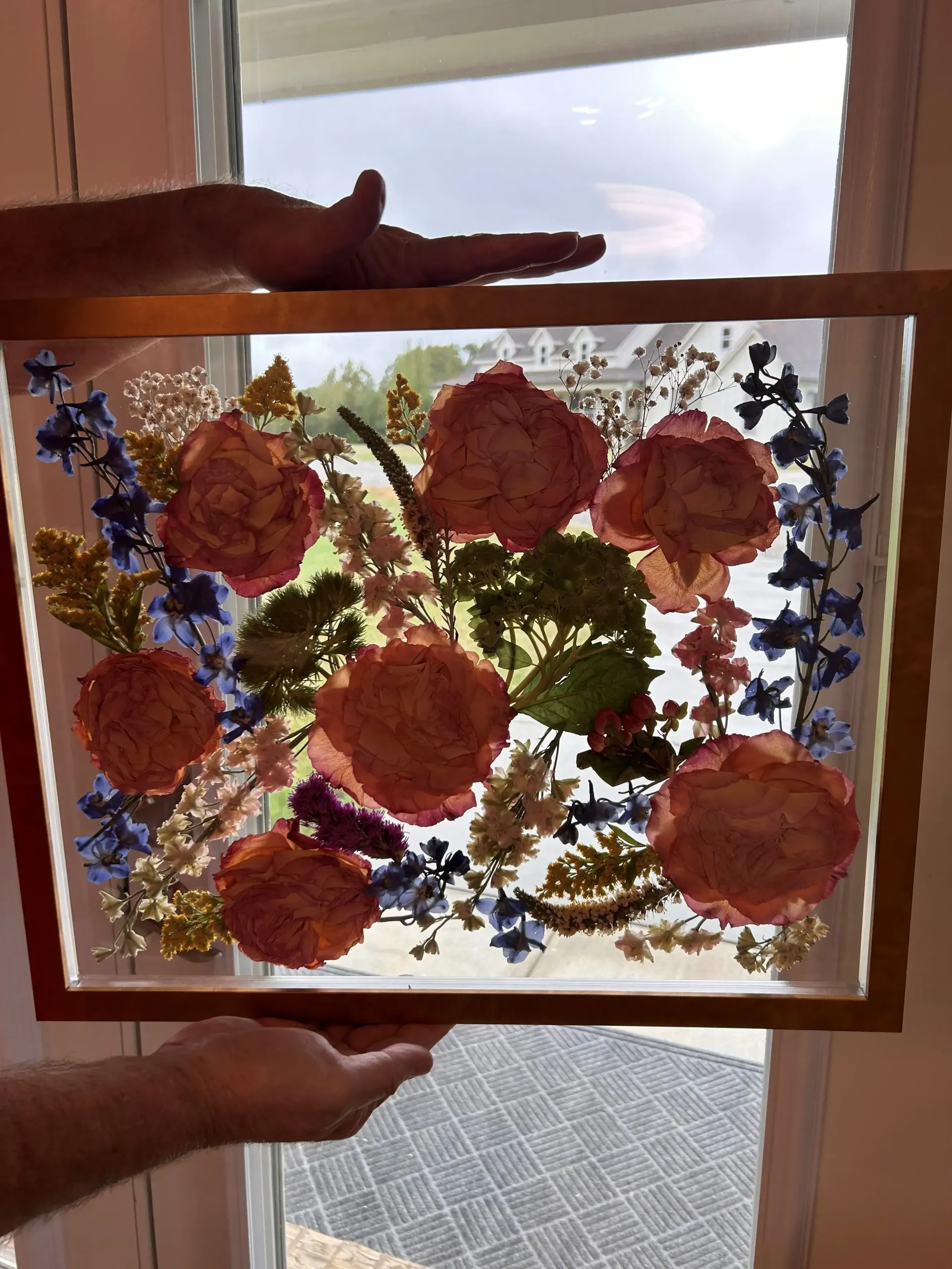 Hands holding a framed stained-glass panel with pink flowers and blue leaves in front of a window