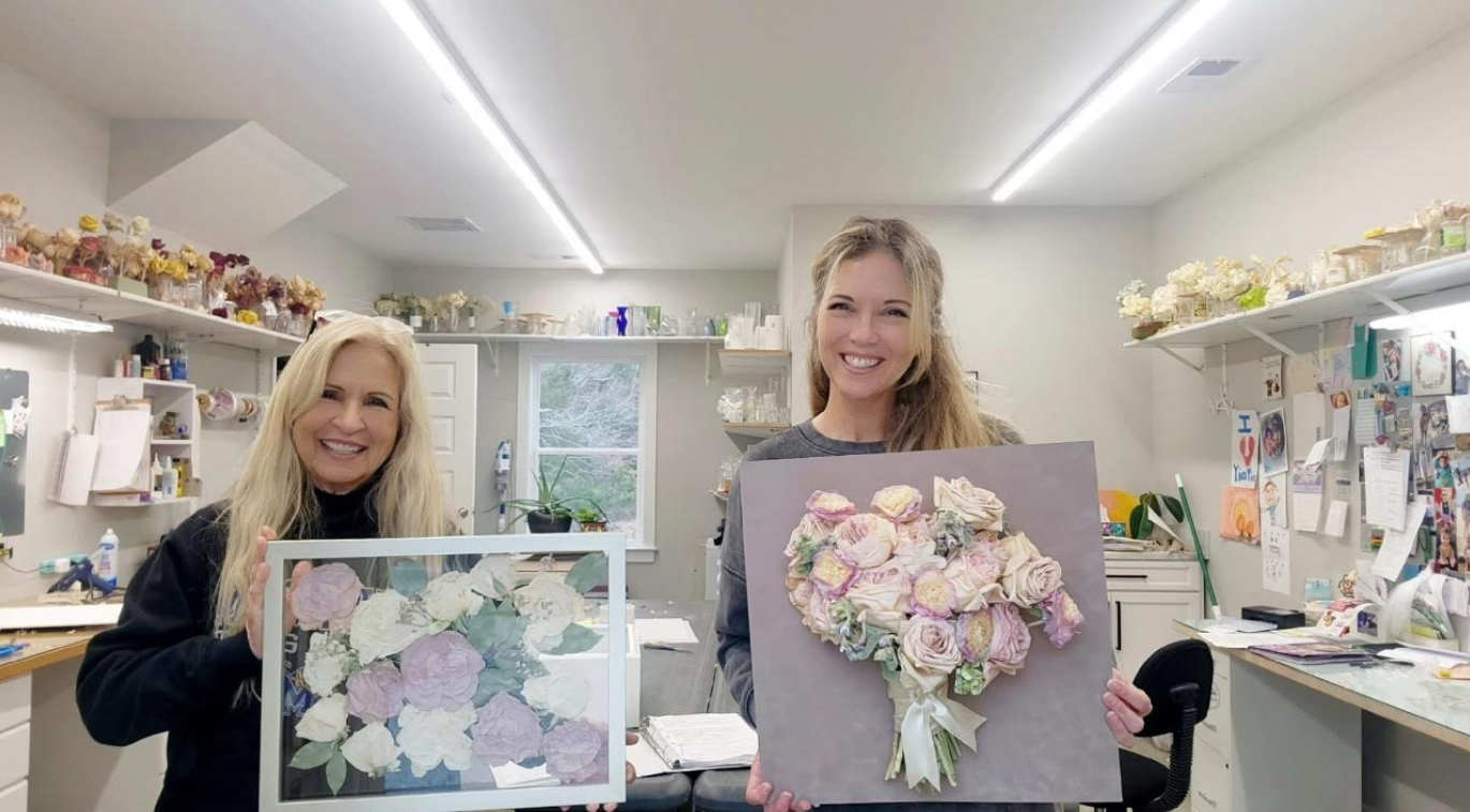 Two women in a craft studio holding framed floral art pieces and smiling