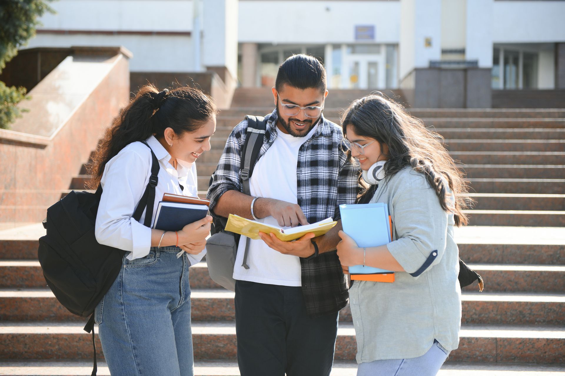 Three students on steps looking at a textbook, smiling. Two women and one man, outdoors in front of a building.