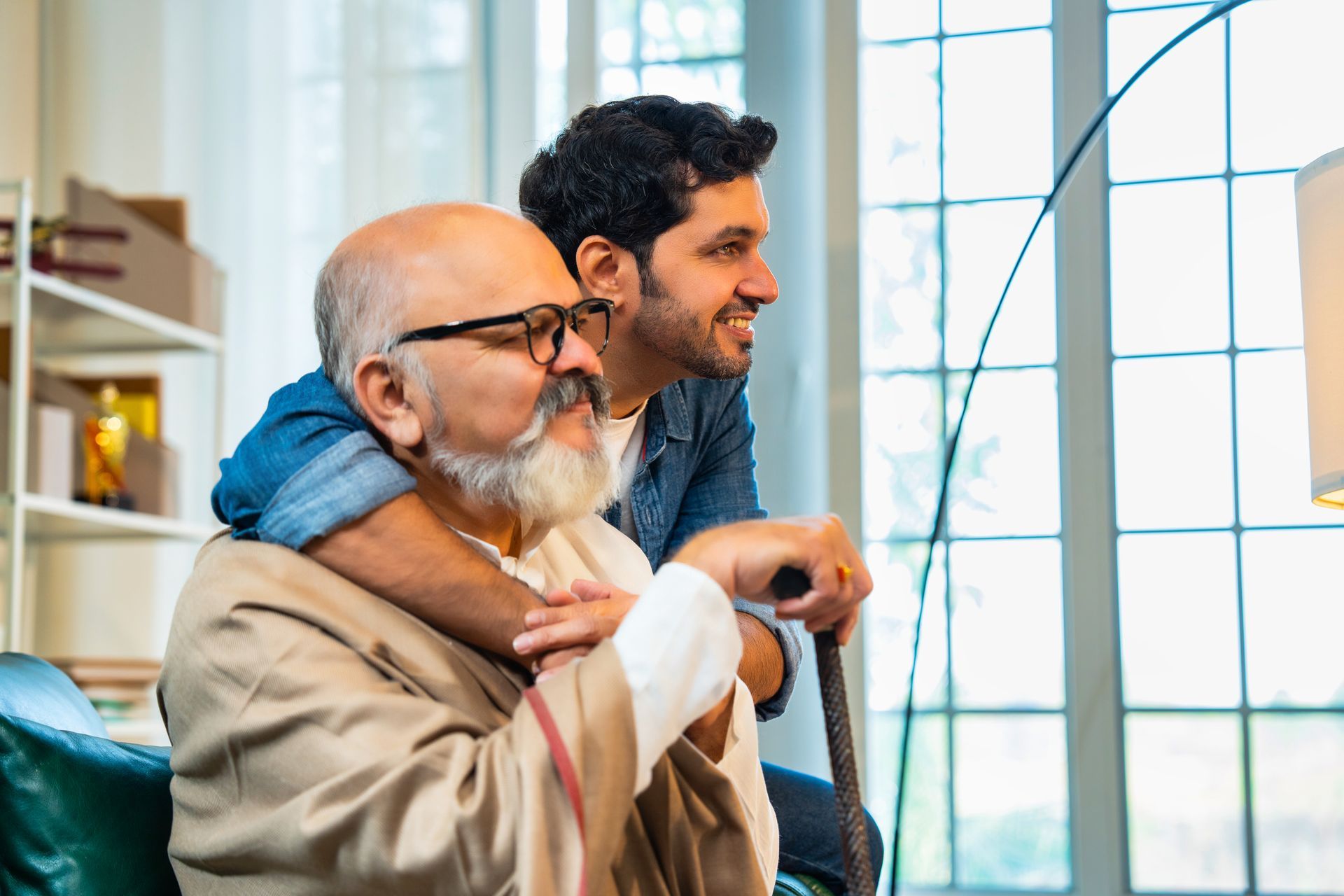 A smiling adult son embraces his elderly father, who leans on a cane and looks out a window, in a living room.