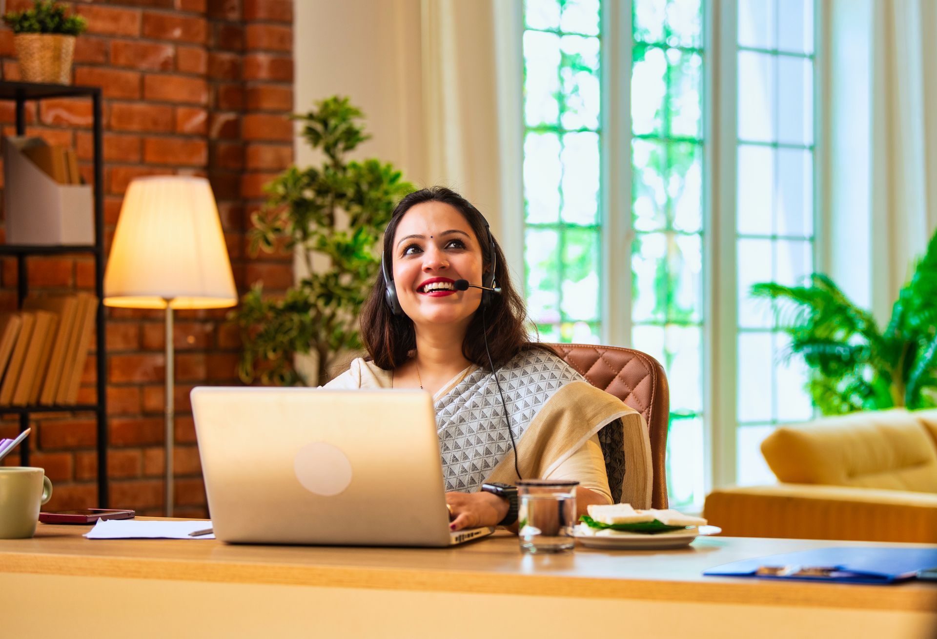 Woman wearing a headset, working on a laptop at a desk, smiling in a home office with a brick wall.
