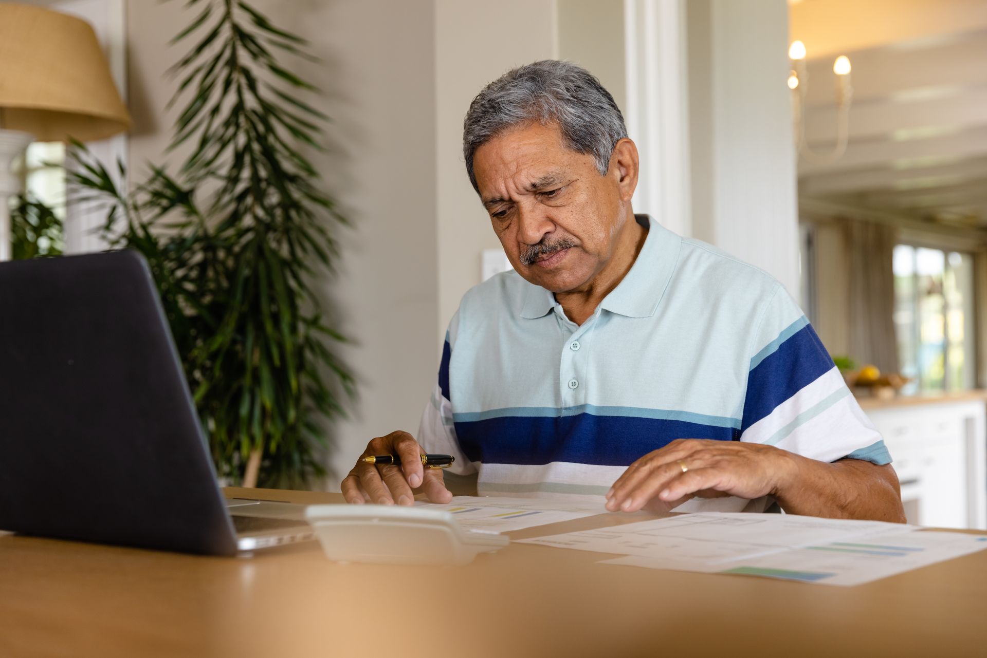 Older man using a calculator and laptop at a table, reviewing documents. He has a focused expression, in a home setting.