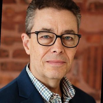 A man with short hair and glasses wearing a blue blazer and plaid shirt against a blurred brick wall background.