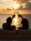 A couple silhouetted against a golden sunset, toasting with champagne glasses while sitting by the ocean.