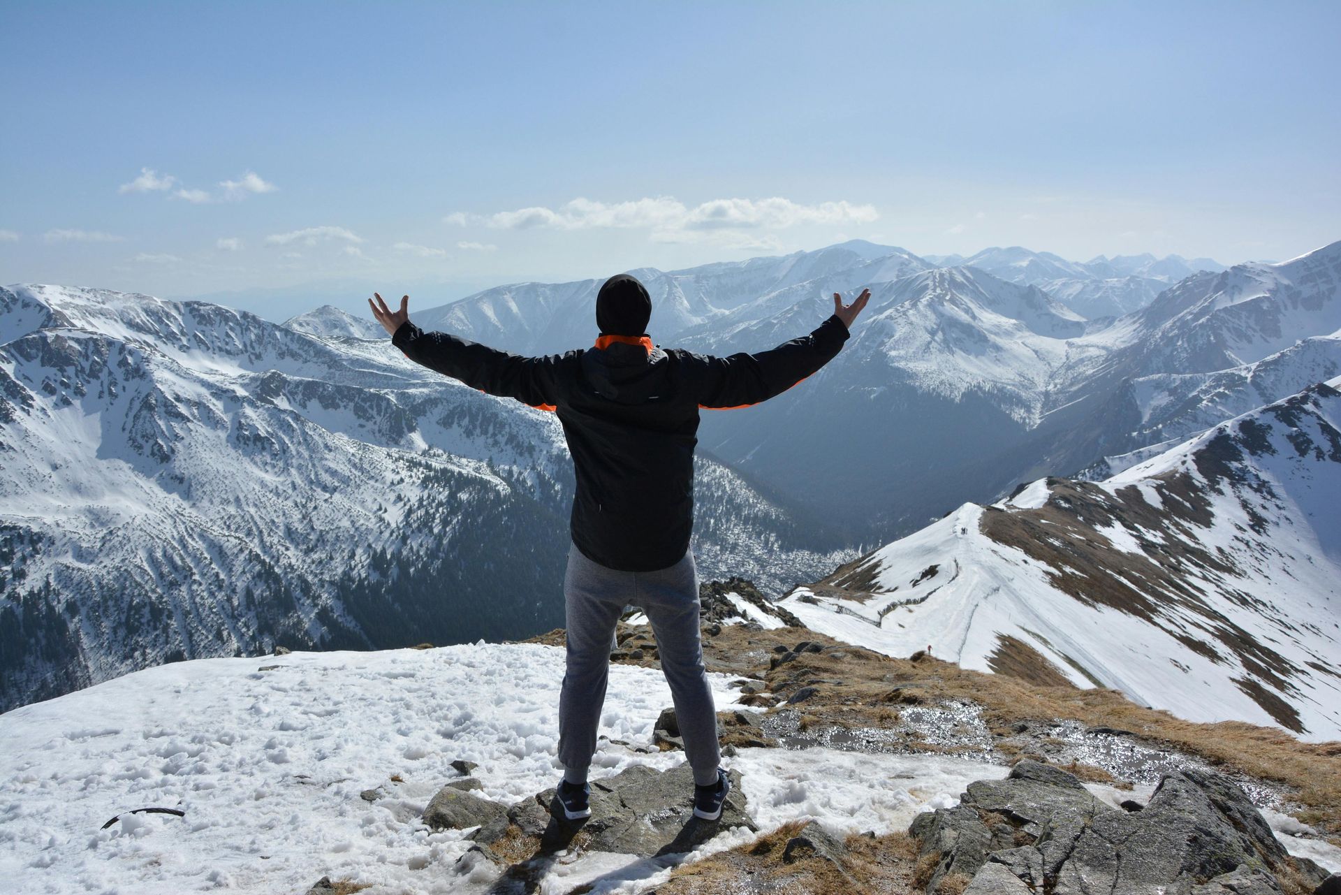 A person stands on a snowy mountain summit with arms outstretched, looking out over a wide range of snow-capped peaks.