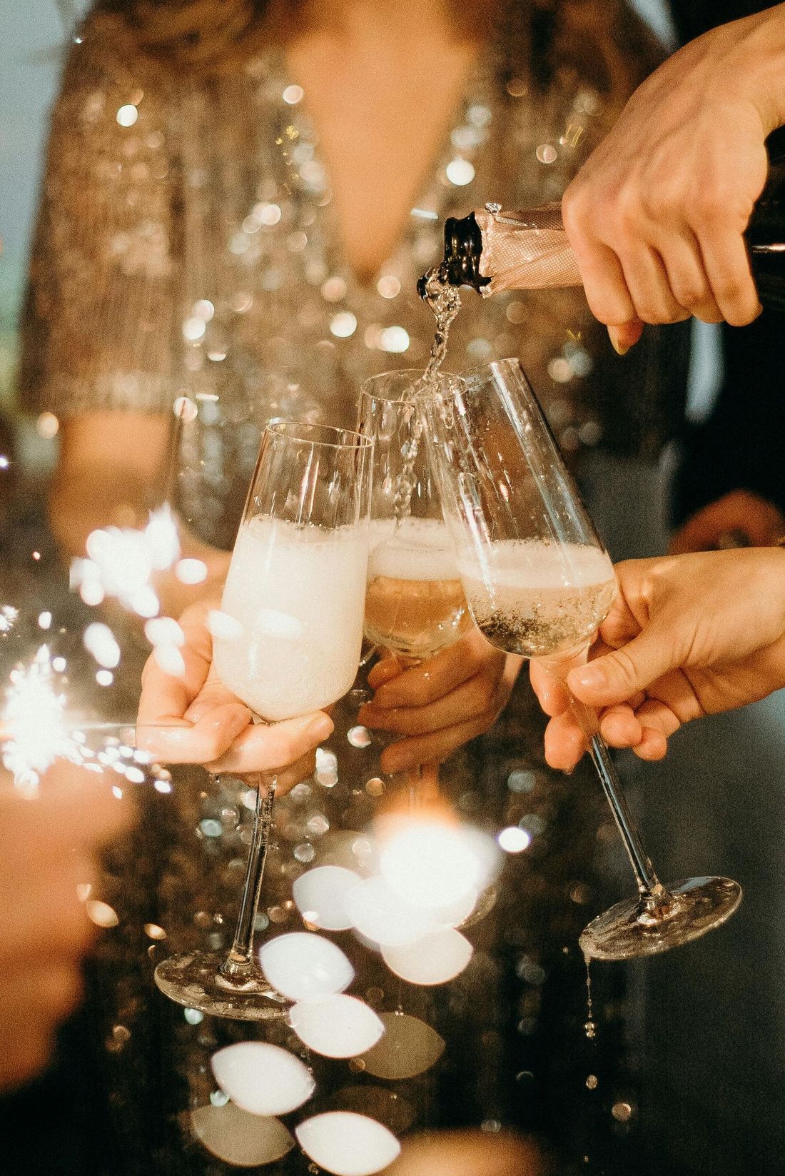 Close-up of hands pouring champagne into three crystal flutes amidst sparkling lights at a festive gathering.