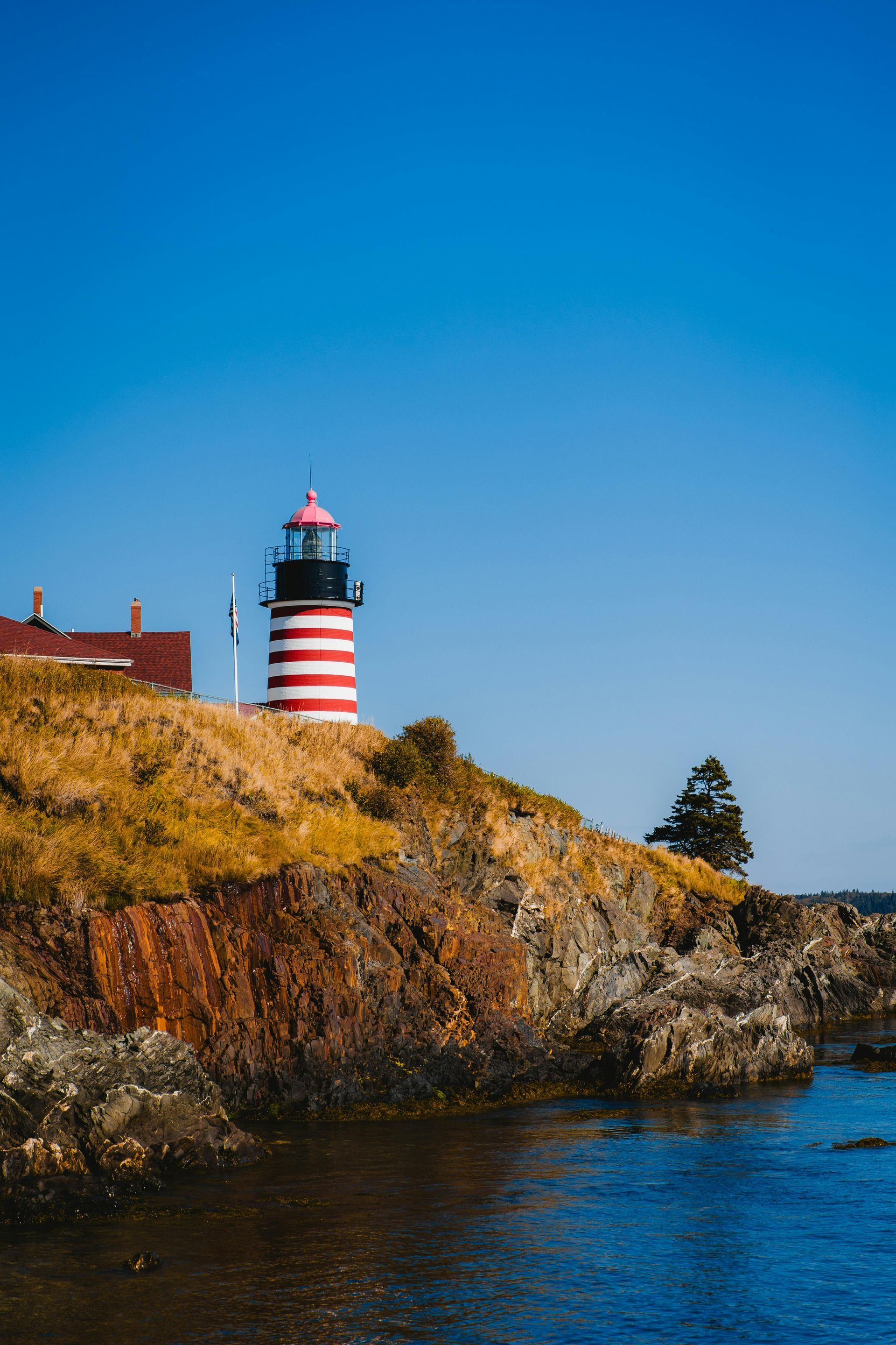 A red and white striped lighthouse stands atop a rocky cliff overlooking the ocean under a clear blue sky.