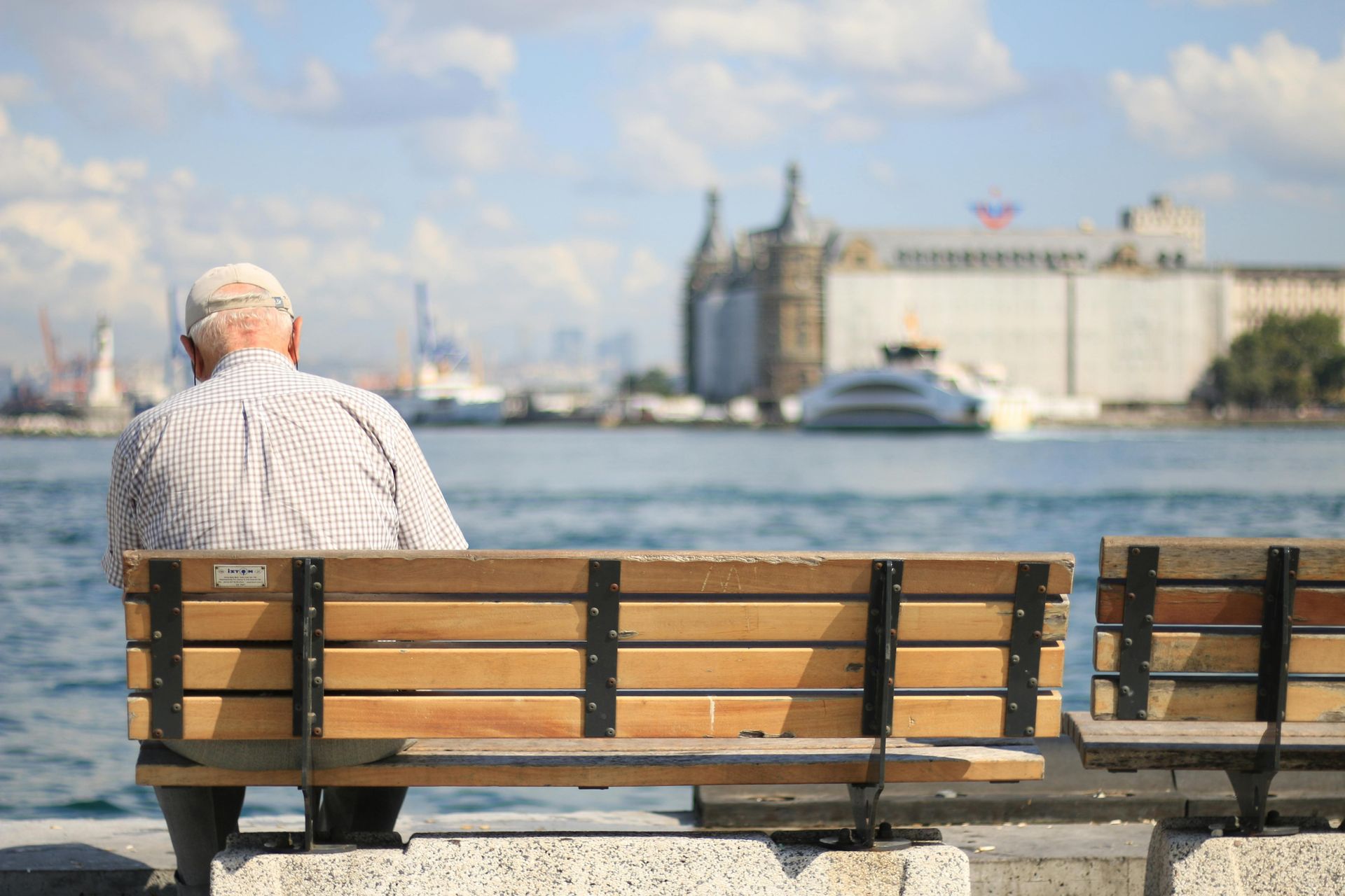 A person sitting on a park bench by the water, looking toward a large building and a boat in the distance.