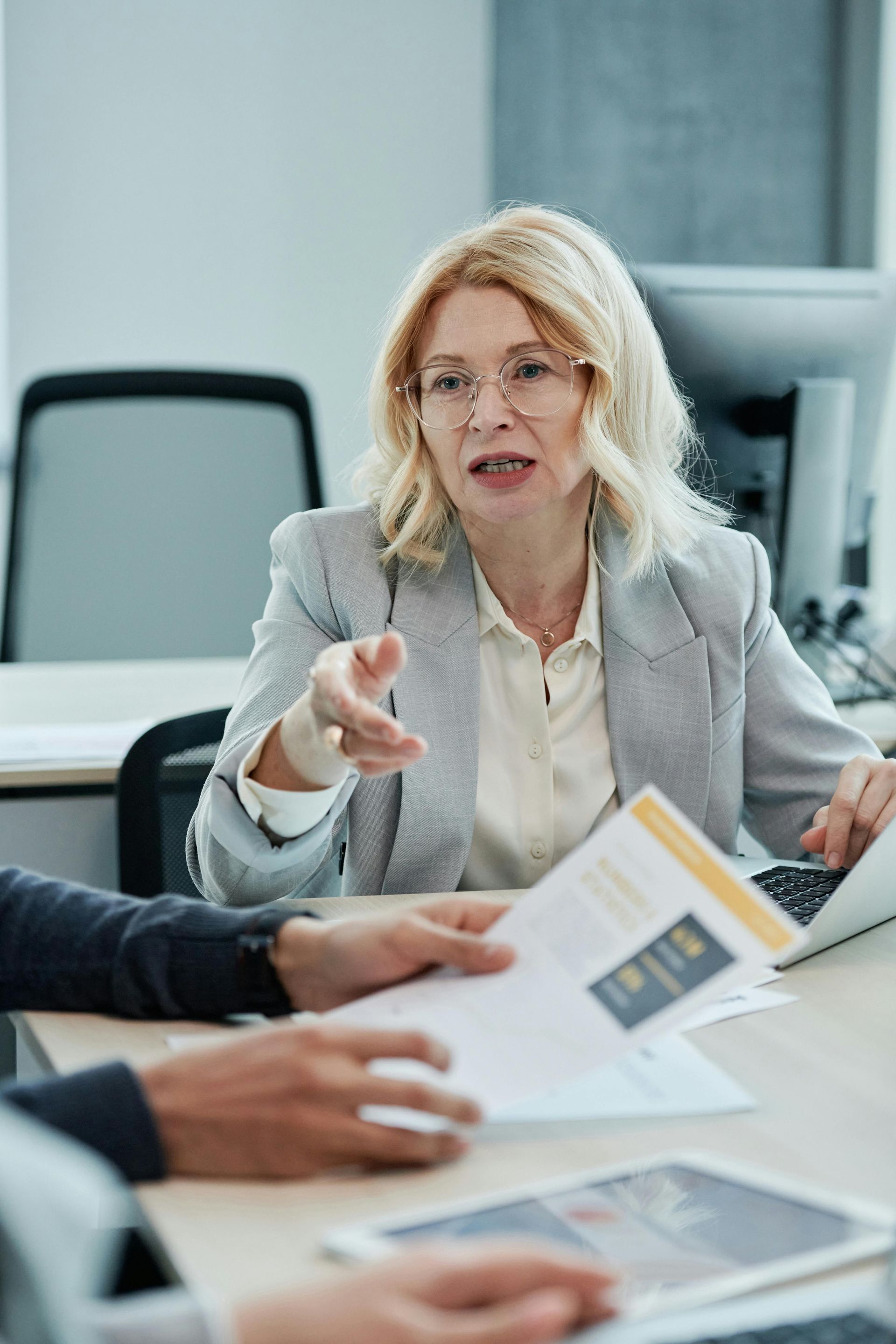 A person in a professional office setting gesturing while speaking to someone holding documents across a desk.