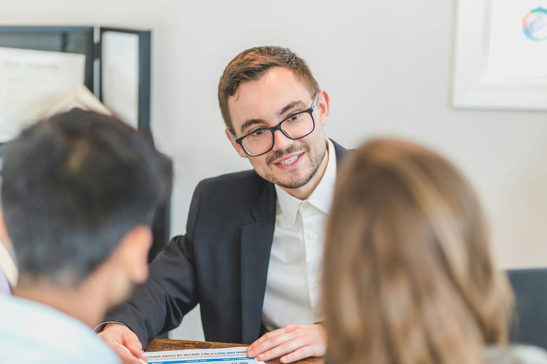 A person in a suit and glasses sits at a table, smiling while looking at two people seated opposite them.