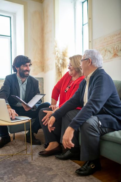 A professional sits with a couple in a brightly lit room, discussing documents on a laptop and in a folder.