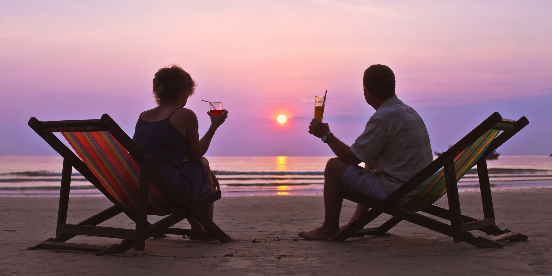 A couple sits in beach chairs on the sand, toasting with drinks while watching a pink and purple sunset over the ocean.
