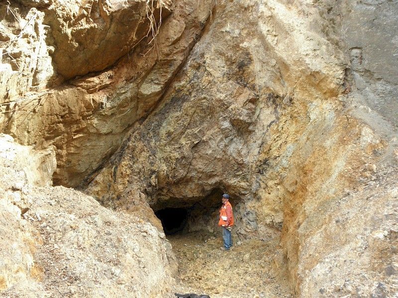 A man in an orange shirt is standing in front of a cave.