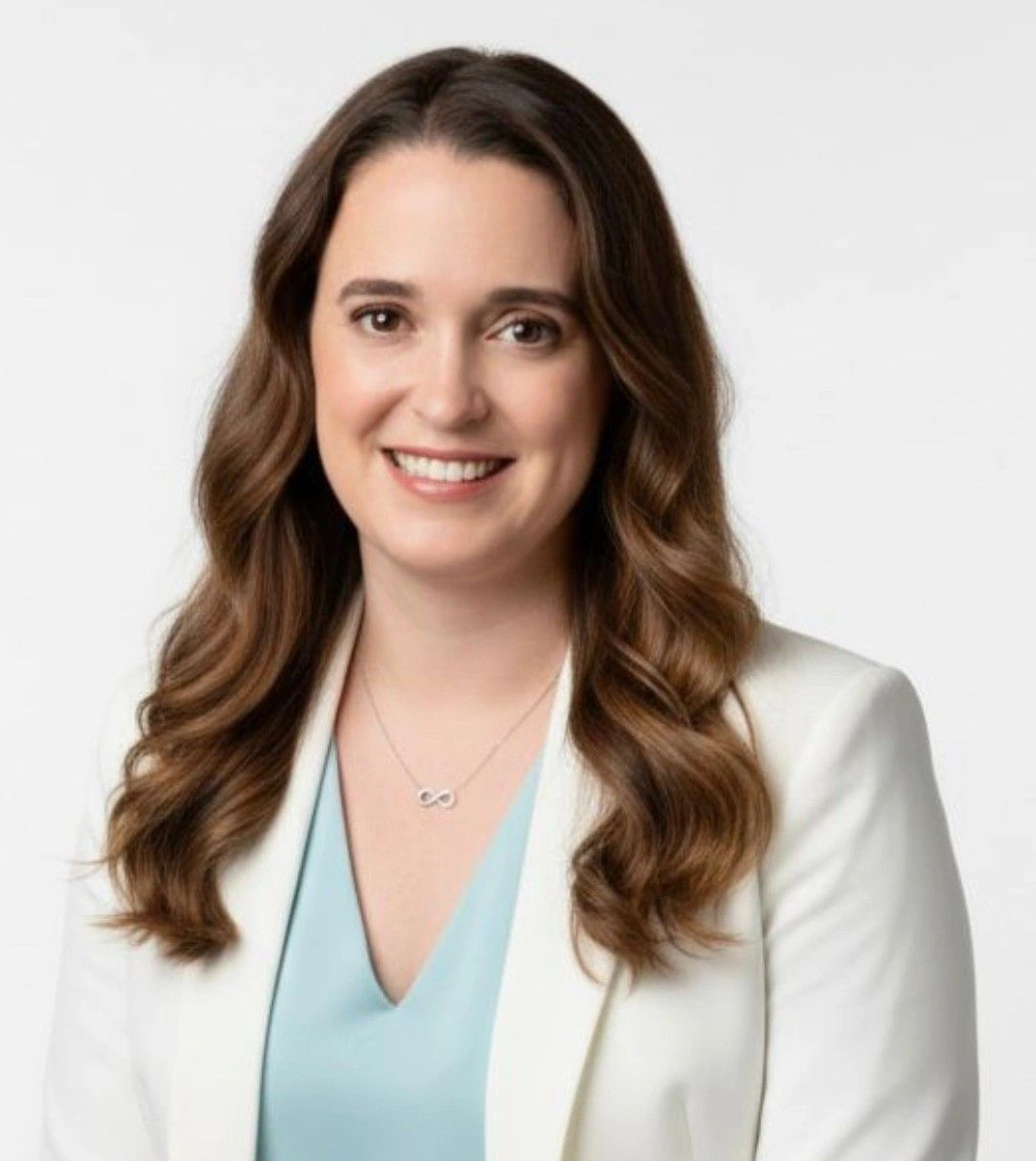 Woman smiling outdoors with brown hair; sunglasses on head; cafe setting.