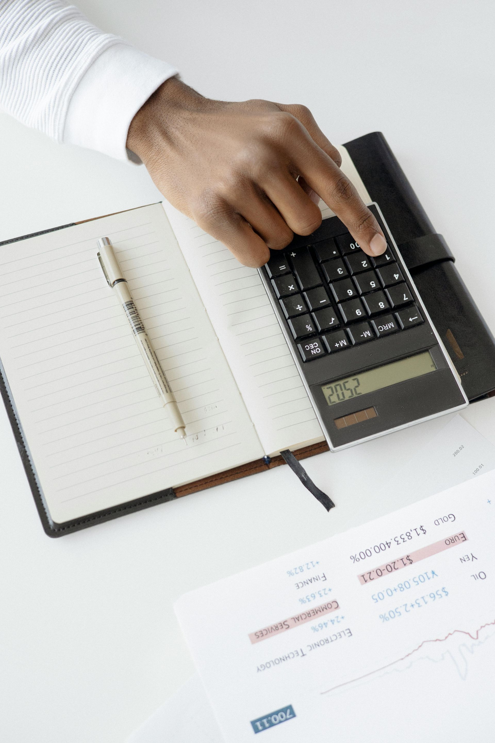 A person is using a calculator next to a notebook and pen.