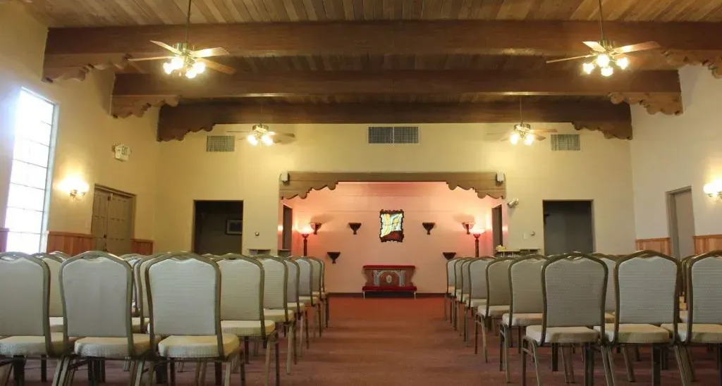 Two people in black suits carry a wooden casket with white flower arrangement.