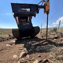 Mini excavator with a post driver working on a fence post in a field.