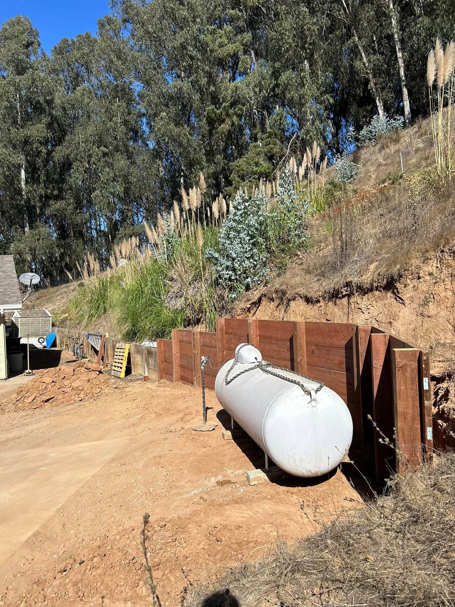 A large, horizontal, white tank next to a retaining wall on a hillside with trees and dirt.