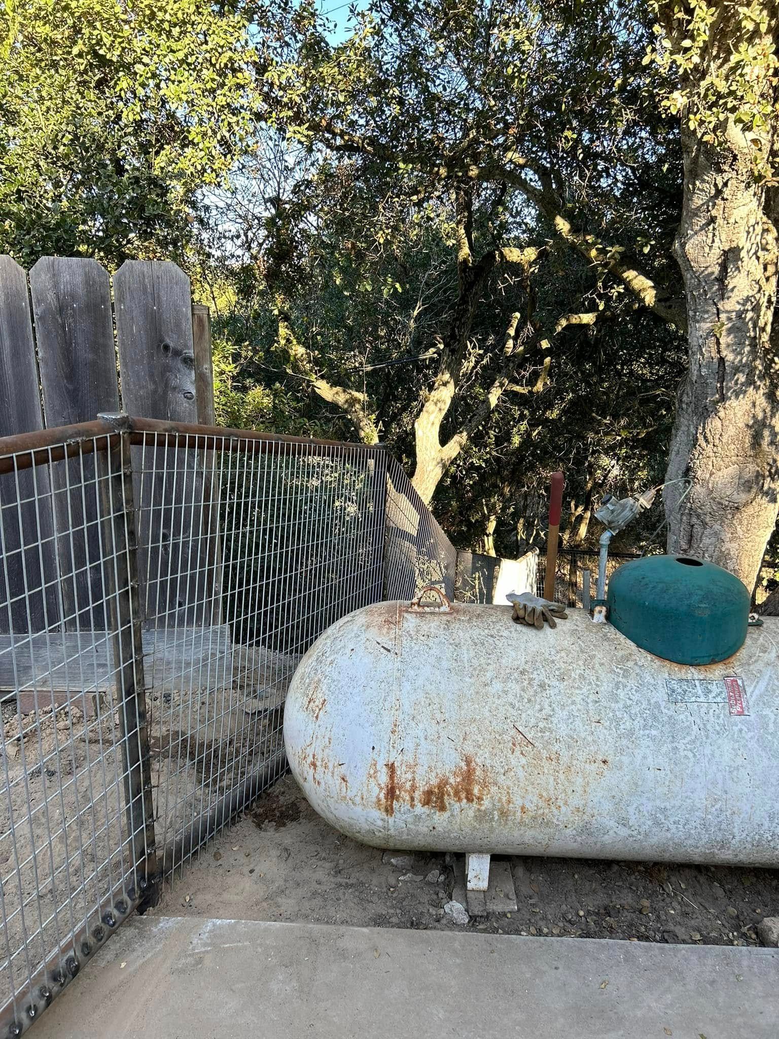 Rusty propane tank next to a chain-link fence, partially obscured by trees.