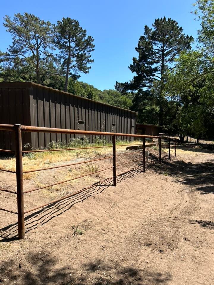 Dark brown building behind a weathered metal fence on a dirt path, with trees in the background under a blue sky.