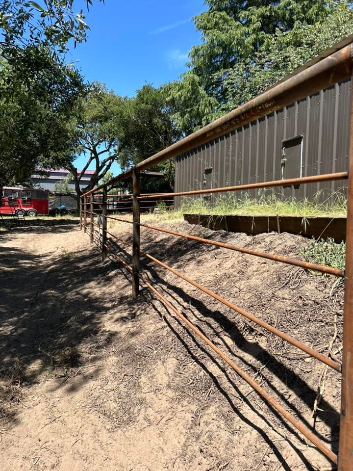 Brown metal fence bordering a dirt path, with a building in the background. Bright blue sky.