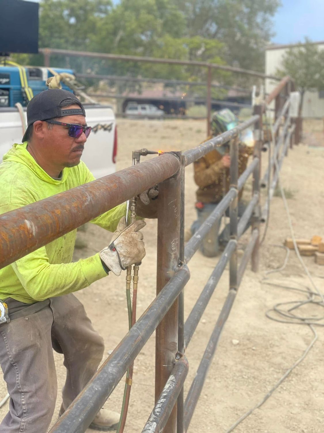 Two workers welding metal fence outdoors; one is using a torch and the other is welding with protective gear.