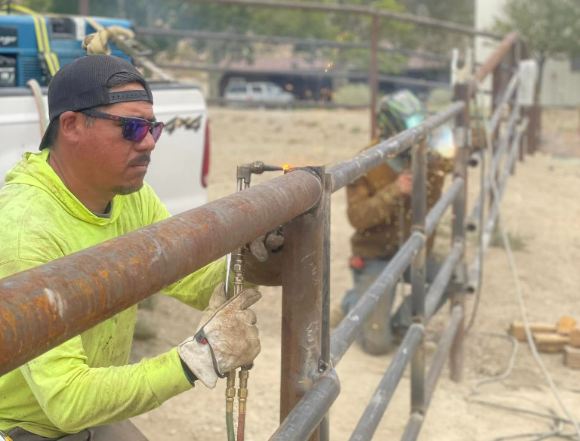 Man in yellow shirt welding metal fence outdoors, sunny day.