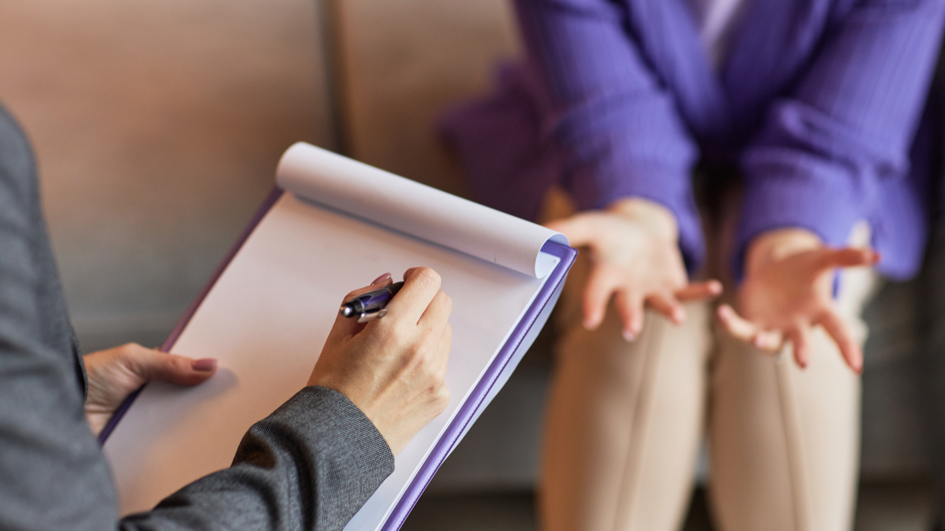 Person in a grey suit takes notes on a clipboard as another person in purple jacket gestures.