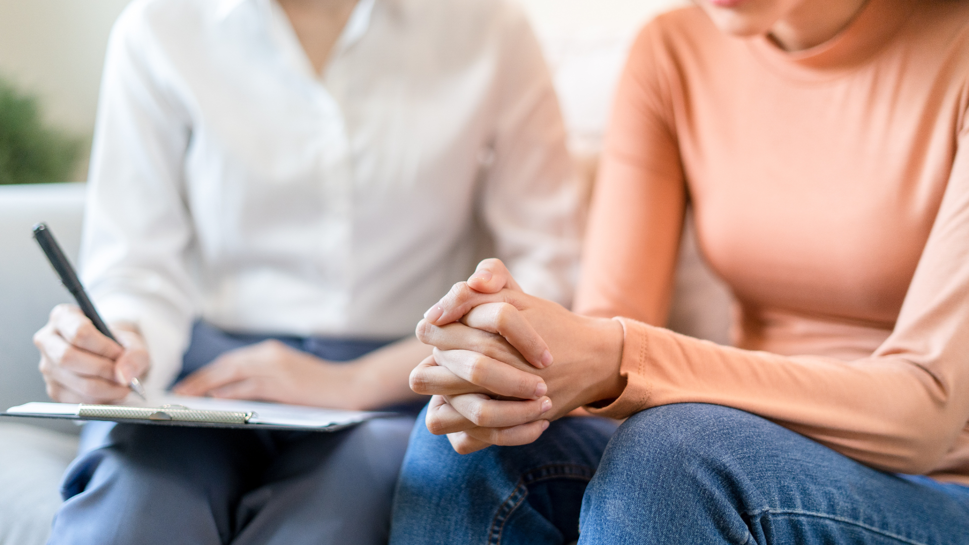 Person in orange shirt with hands clasped, sitting with someone taking notes.